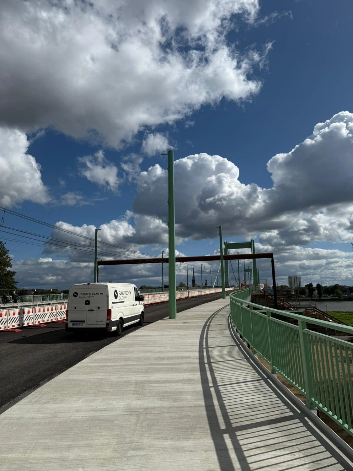 Fugentechnik Buchen - Bau - Ein weißer Lieferwagen parkt auf einer Brücke mit grünen Geländern und Laternenpfählen unter einem teilweise bewölkten blauen Himmel