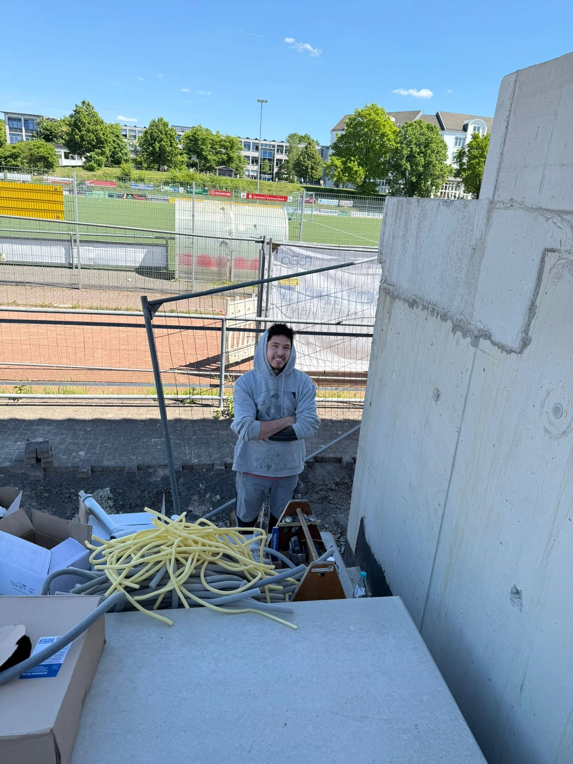 Fugentechnik Buchen - Bau - Mann im grauen Kapuzenpullover steht auf einer Baustelle mit aufgerollten gelben und grauen Kabeln in der Nähe und einem Sportplatz im Hintergrund unter klarem blauen Himmel