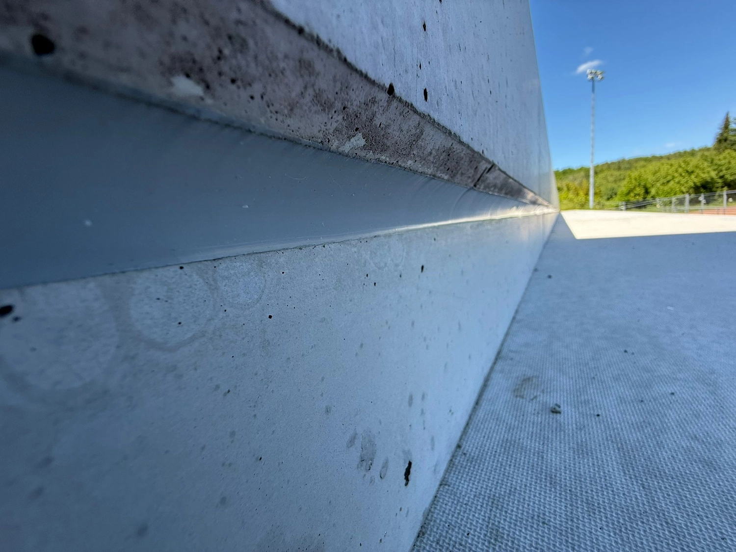 Fugentechnik Buchen - Bau - Nahaufnahme einer Betonwand und einer strukturierten Bodenfläche, die sich in die Ferne erstreckt, mit Bäumen und einem Lichtmast unter blauem Himmel