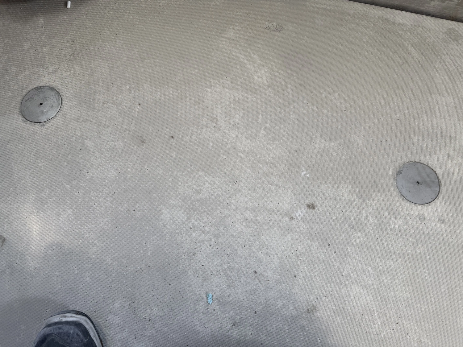 Fugentechnik Buchen - Bau - Hellgraue, strukturierte Oberfläche mit zwei runden Metall-Ablaufabdeckungen und einem teilweise sichtbaren Schuh in der unteren linken Ecke
