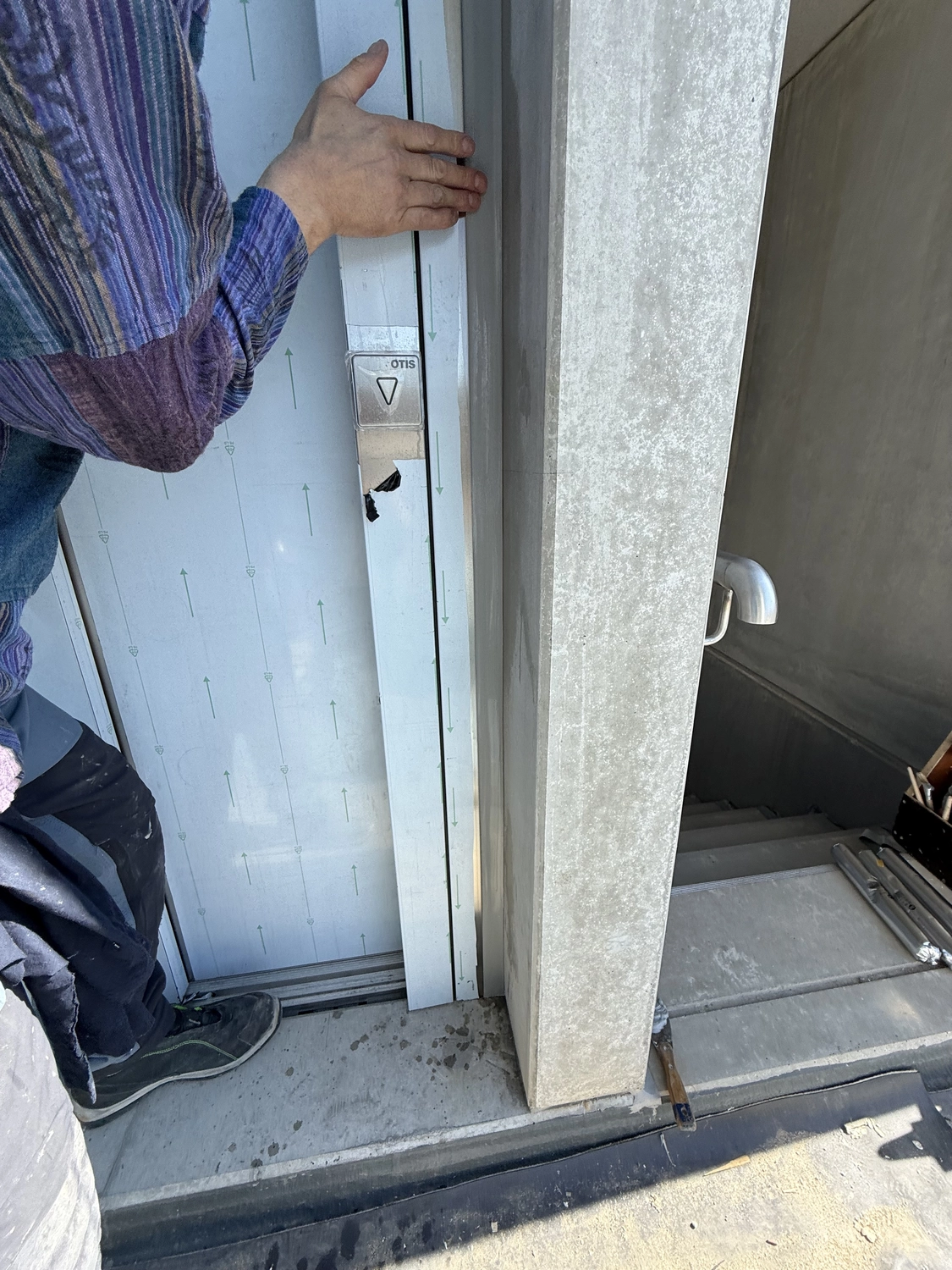 Fugentechnik Buchen - Bau - Person drückt in einem Betontreppenhaus eine teilweise geöffnete Aufzugstür mit beschädigtem Bedienfeld auf.
