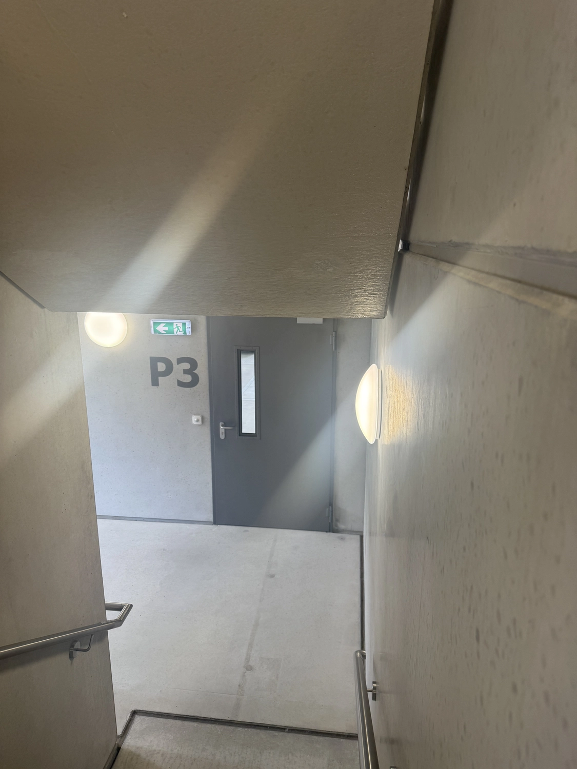 Fugentechnik Buchen - Bau - Betontreppenhaus, das zu einem Flur mit einer grauen Tür mit der Aufschrift P3 und einem darüber befindlichen Notausgangsschild führt