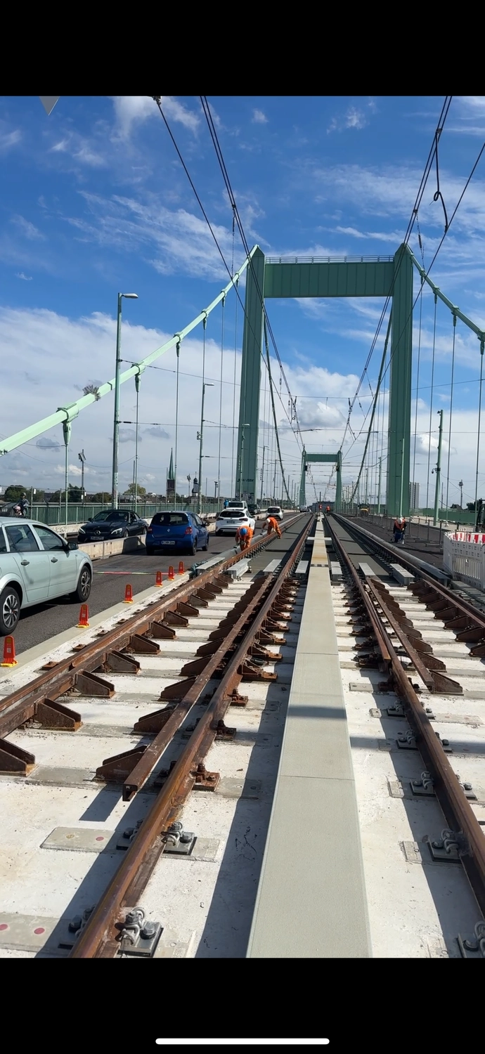 Fugentechnik Buchen - Bau - Arbeiter in orangefarbener Schutzkleidung arbeiten an den Gleisen einer Hängebrücke, während auf den angrenzenden Fahrspuren Autos unter blauem Himmel fahren