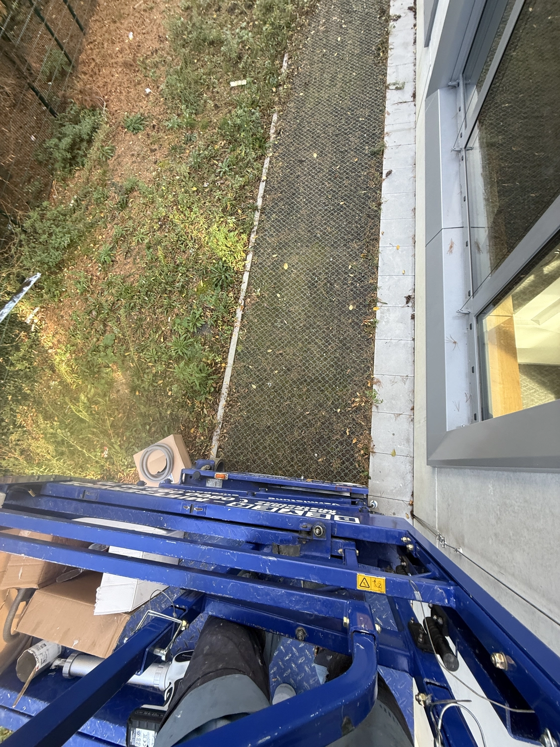 Fugentechnik Buchen - Bau - Blick von einer erhöhten blauen hydraulischen Hebebühne neben einem Gebäude mit Glastüren nach unten, auf einen schmalen Gehweg, Gras und verstreute Blätter unterhalb