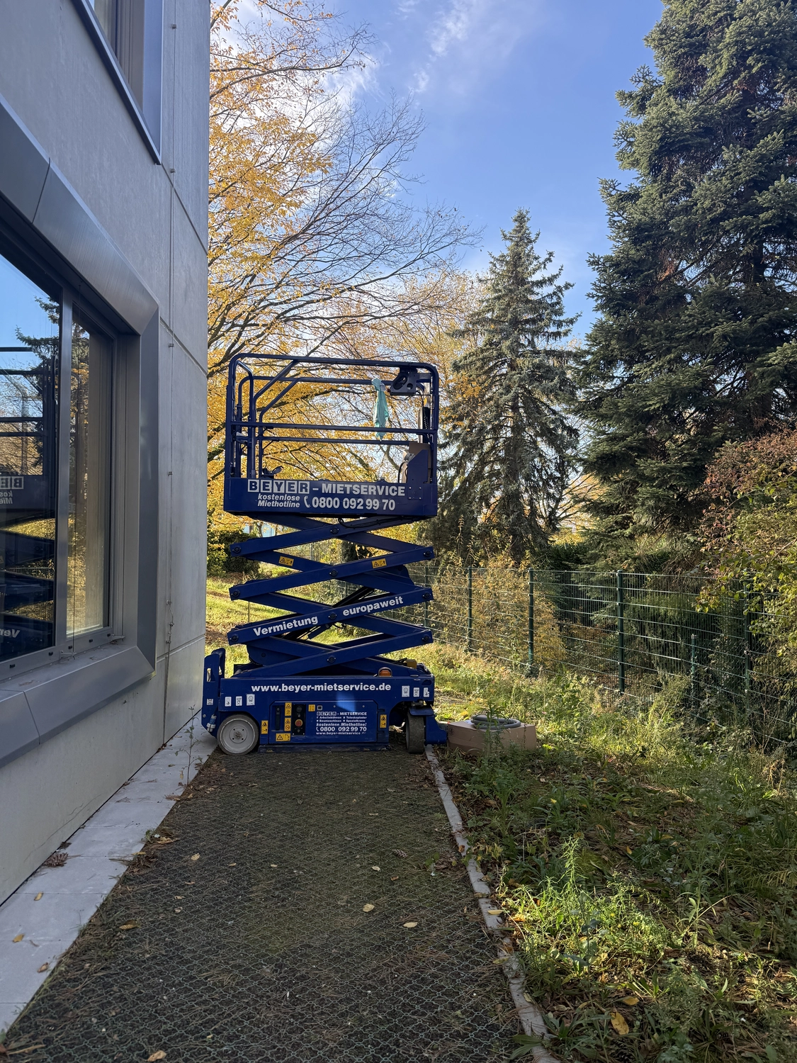 Fugentechnik Buchen - Bau - Blaue Scherenbühne, geparkt auf einem schmalen Weg neben einem Gebäude, rechts davon Herbstbäume und ein Drahtzaun