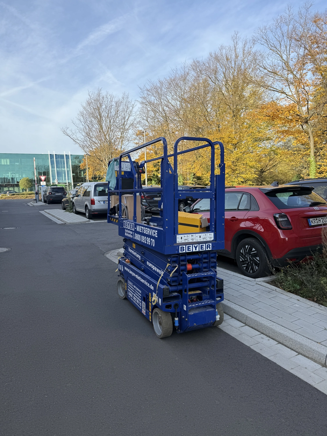 Fugentechnik Buchen - Bau - Blue BEYER Miet-Scherenbühne, geparkt auf einer Straße neben Autos und Herbstbäumen