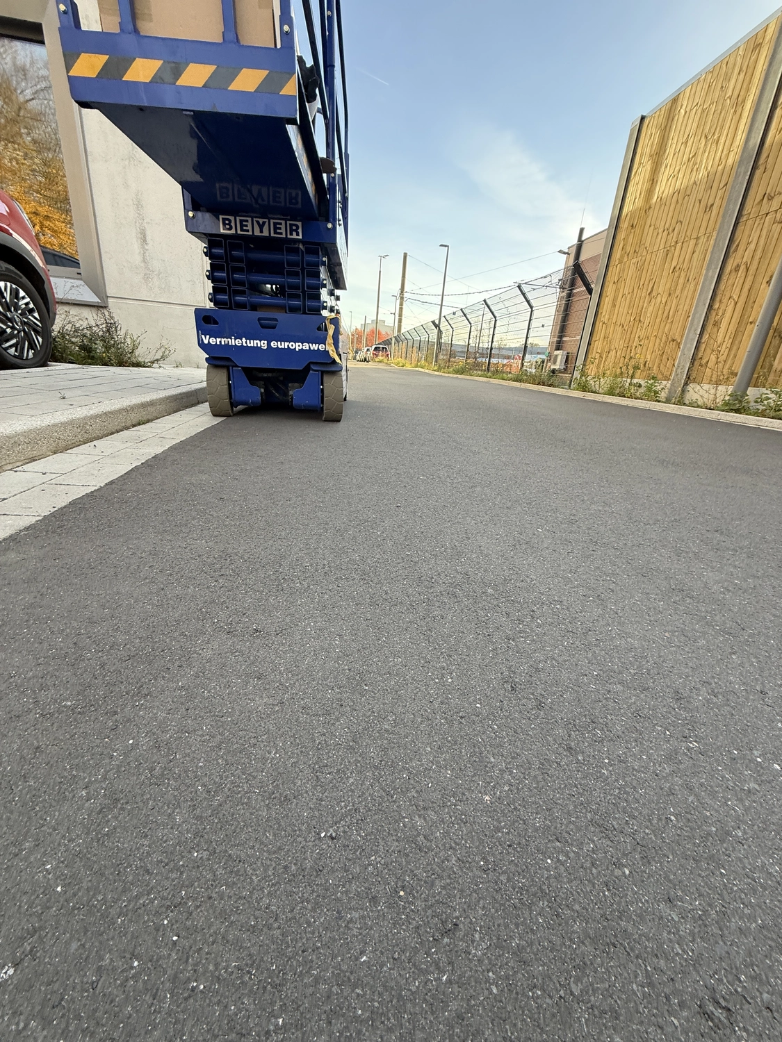 Fugentechnik Buchen - Bau - Nahaufnahme einer blauen Industriehebebühne mit der Aufschrift „BEYER“ auf einer Asphaltstraße zwischen einem Gebäude mit Stacheldrahtzaun und einem Holzzaun