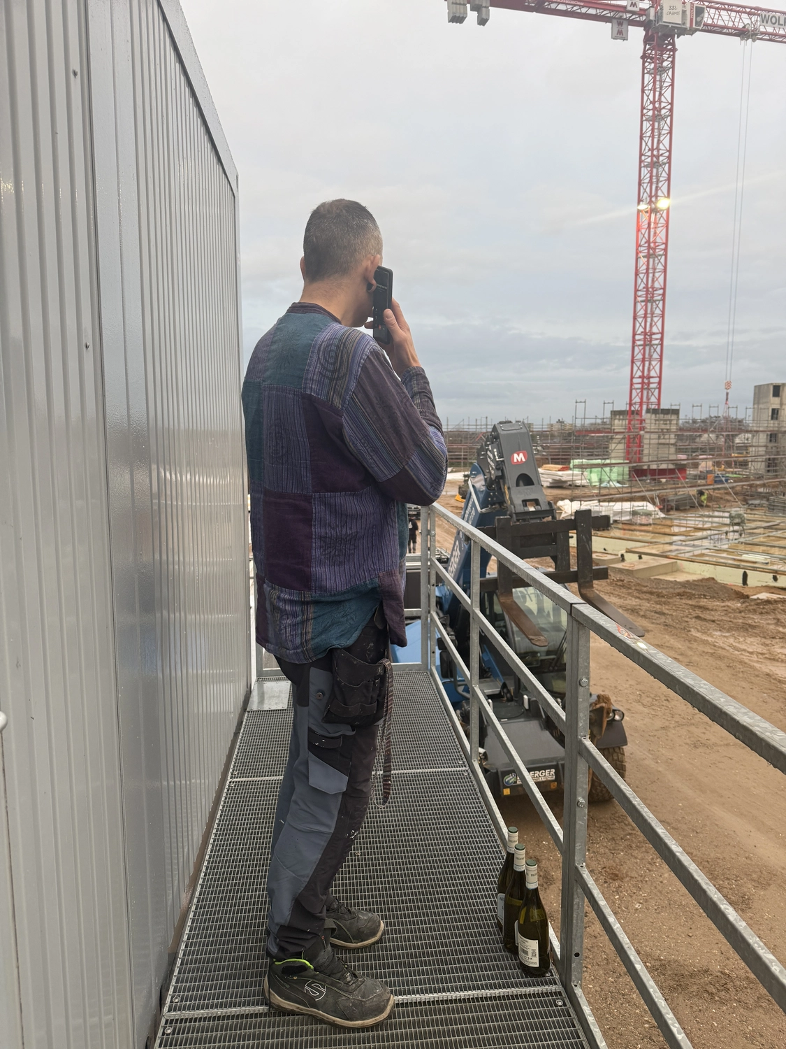 Fugentechnik Buchen - Bau - Ein Mann in Arbeitskleidung telefoniert auf einer Metallplattform mit Blick auf eine Baustelle mit Kran und Baumaschinen.