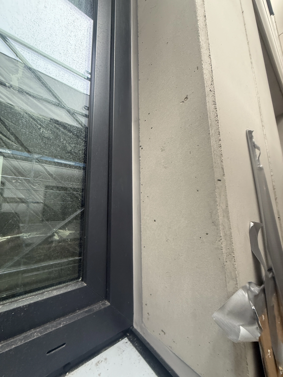 Fugentechnik Buchen - Bau - Nahaufnahme eines Eckfensterrahmens an einem Gebäude mit reflektierender Glasscheibe und beiger Wand, an der etwas Schmutz und sich ablösendes Klebeband zu sehen sind.
