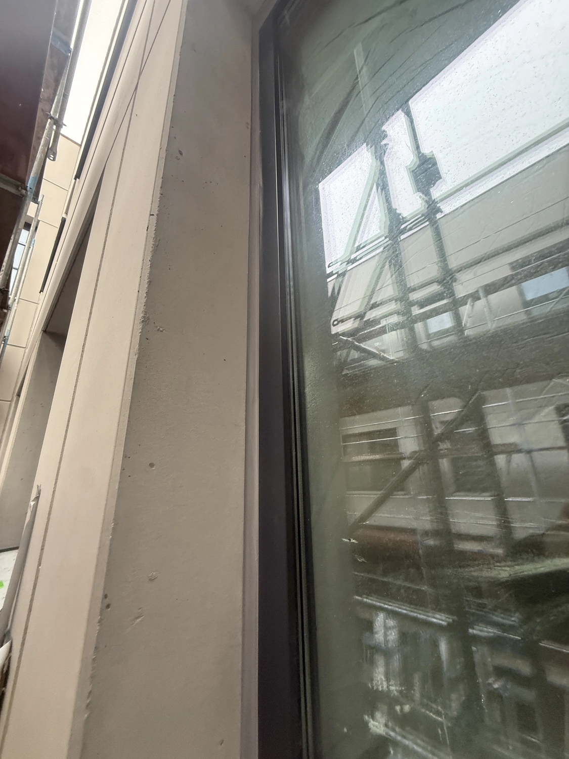 Fugentechnik Buchen - Bau - Nahaufnahme einer Gebäudefassade mit einem Fenster mit Metallrahmen und der Spiegelung eines Baugerüsts im Glas