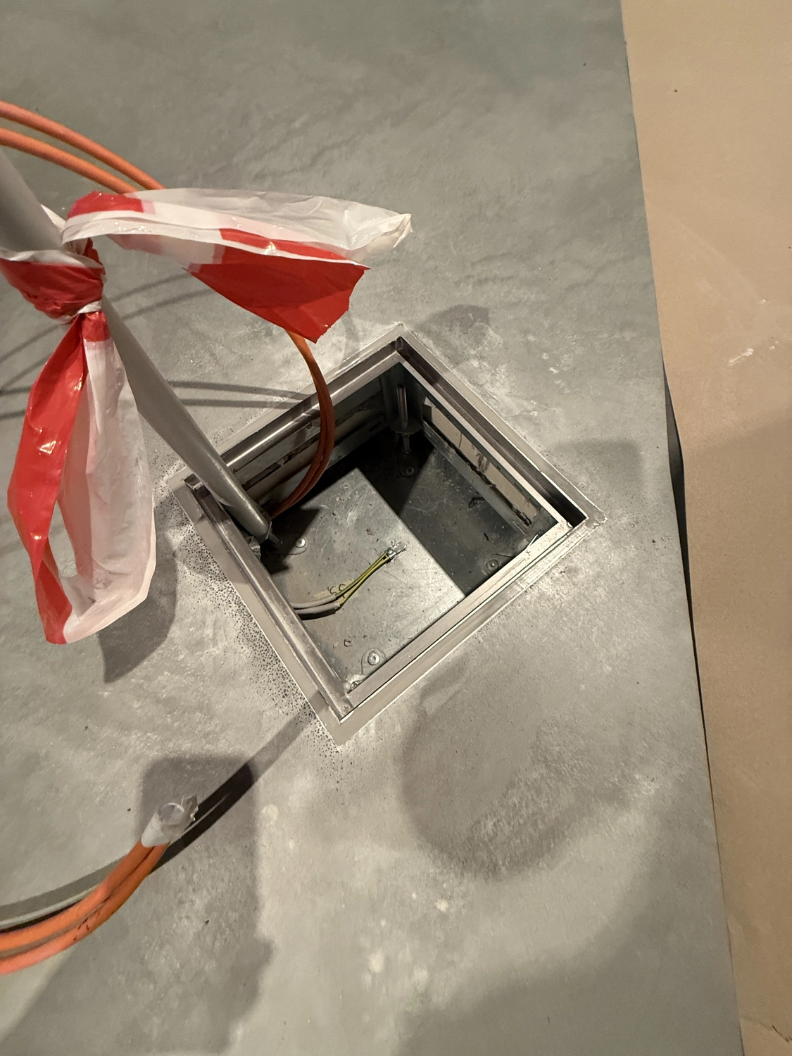 Fugentechnik Buchen - Bau - Offener Bodenverteilerkasten in einem Betonboden mit orangefarbenen Kabeln und einem mit rot-weißem Absperrband umwickelten Mast