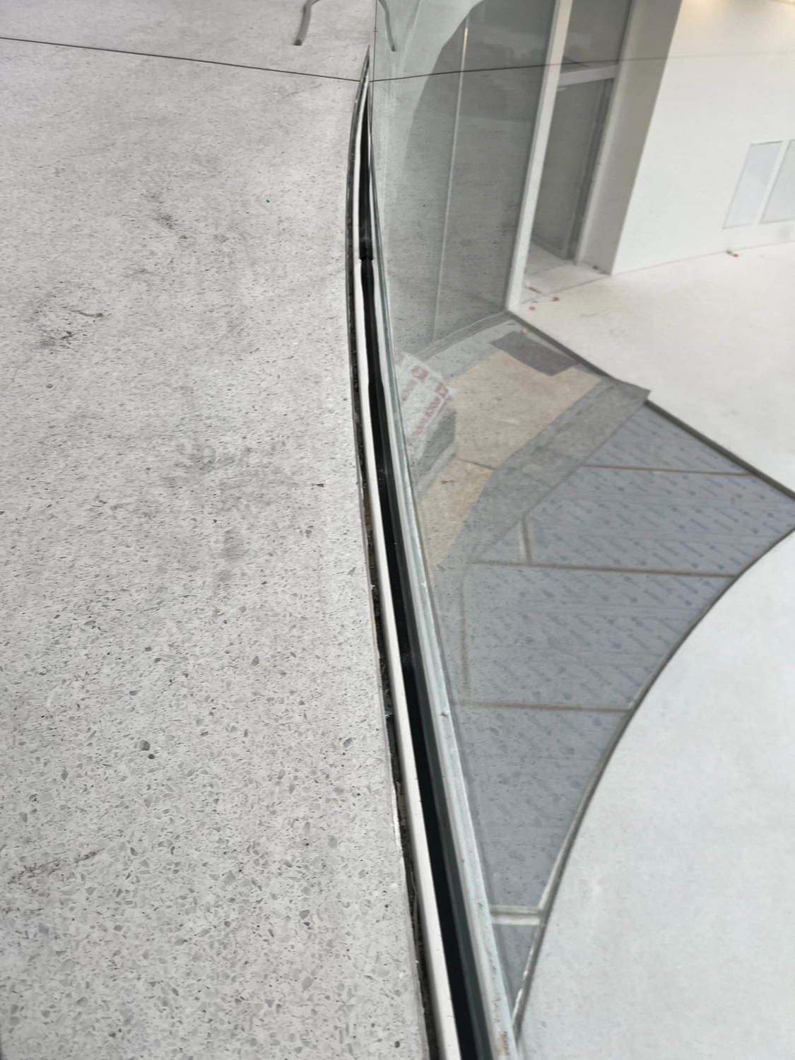 Fugentechnik Buchen - Bau - Nahaufnahme eines schmalen Spalts zwischen einer Betonfläche und einer Glaswand, in der sich ein gefliester Boden spiegelt.