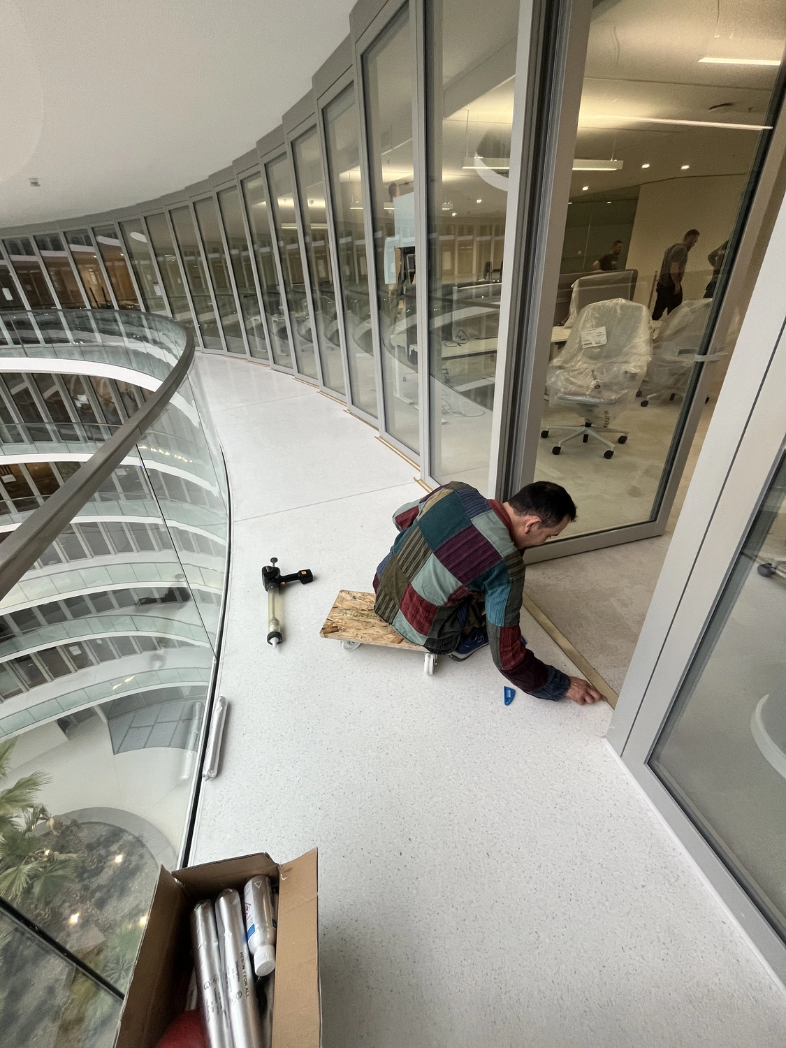 Fugentechnik Buchen - Bau - Mann mit buntem Patchworkhemd, brauner Hose mit Farbflecken und blauen Schuhen auf einem Innenbalkon mit Blick auf ein mehrstöckiges Atrium mit Glasgeländer