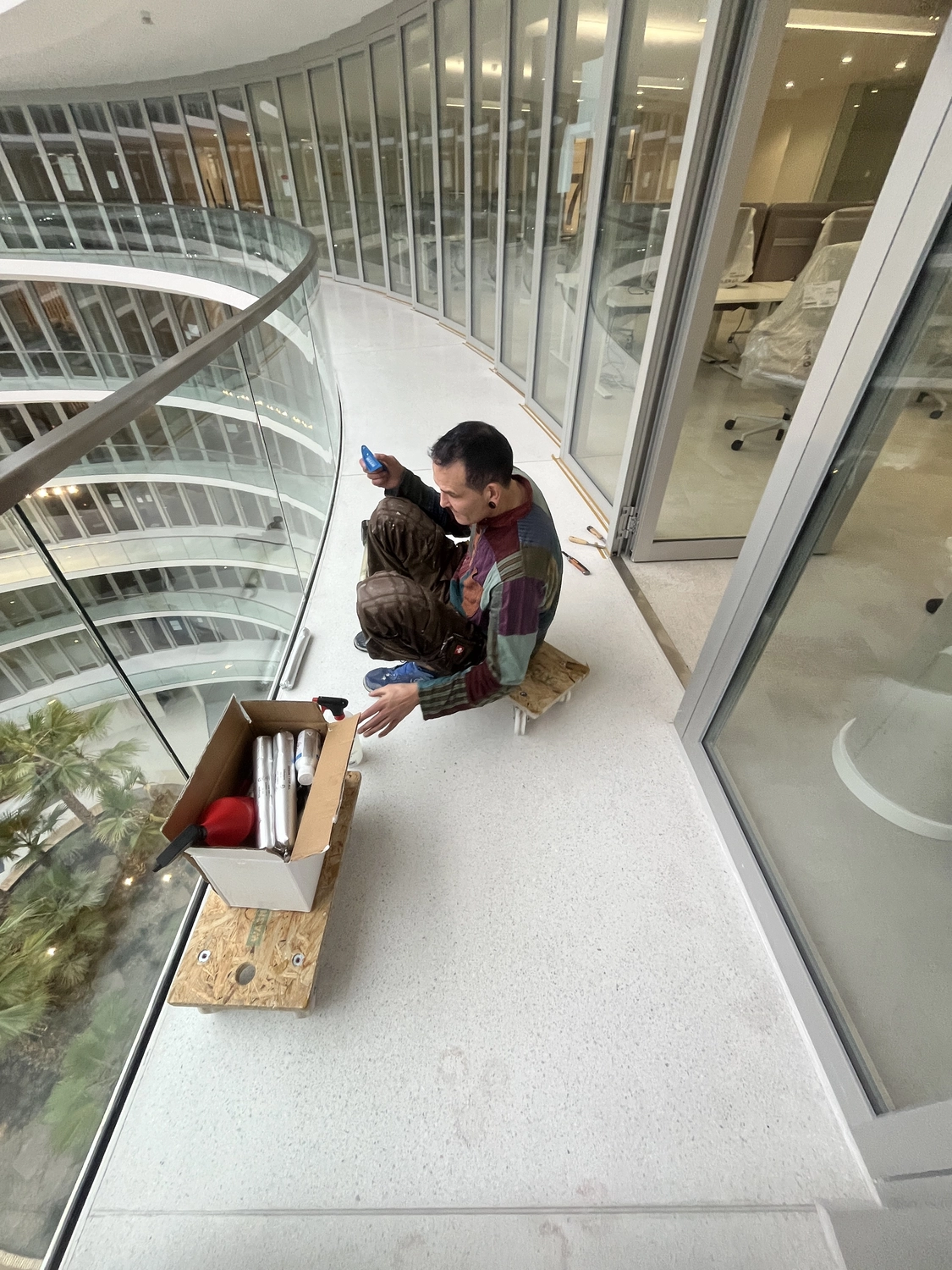 Fugentechnik Buchen - Bau - Mann mit buntem Patchworkhemd, brauner Hose mit Farbflecken und blauen Schuhen auf einem Innenbalkon mit Blick auf ein mehrstöckiges Atrium mit Glasgeländer