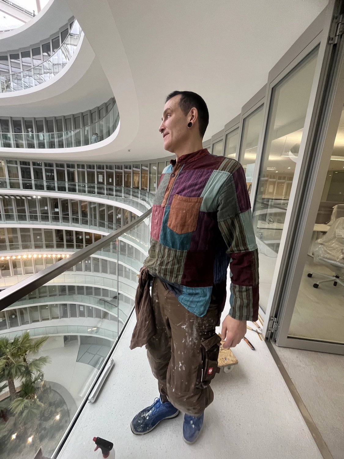 Fugentechnik Buchen - Bau - Mann mit buntem Patchworkhemd, brauner Hose mit Farbflecken und blauen Schuhen steht auf einem Innenbalkon mit Blick auf ein mehrstöckiges Atrium mit Glasgeländer