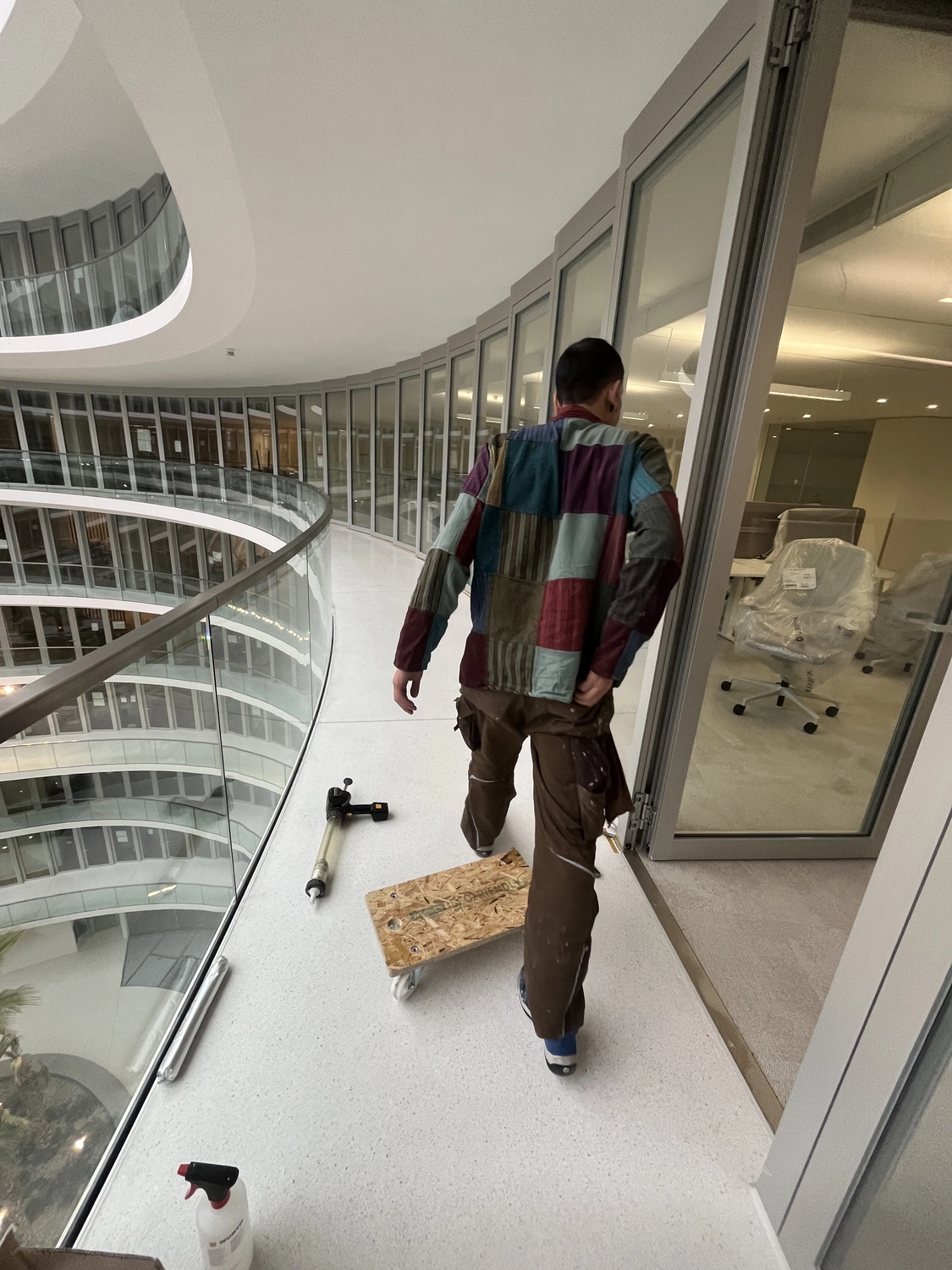 Fugentechnik Buchen - Bau - Mann mit buntem Patchworkhemd, brauner Hose mit Farbflecken und blauen Schuhen auf einem Innenbalkon mit Blick auf ein mehrstöckiges Atrium mit Glasgeländer