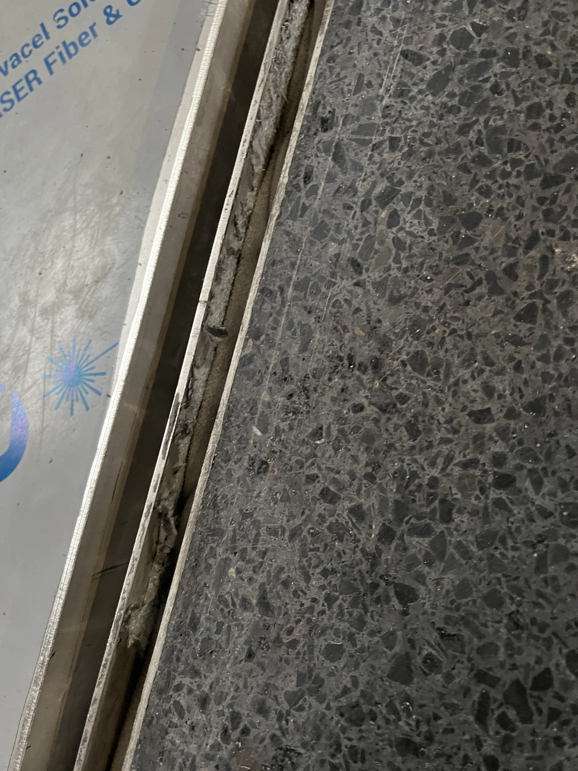 Fugentechnik Buchen - Bau - Nahaufnahme einer rissigen und abgenutzten Bodenfuge zwischen einer gesprenkelten dunkelgrauen Fliese und einer glatten Oberfläche mit teilweise blauem Text und einer sternförmigen Grafik.