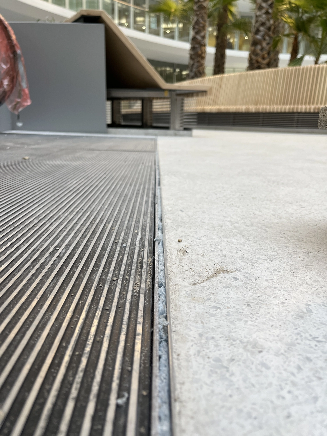 Fugentechnik Buchen - Bau - Nahaufnahme eines strukturierten Metallgitters neben glattem Betonpflaster mit einer Bank und Palmen im Hintergrund