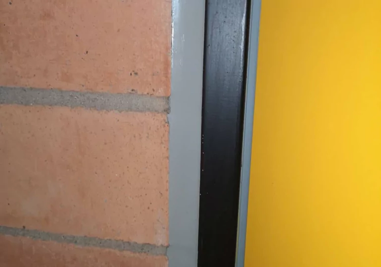 Fugentechnik Buchen - Bau - Nahaufnahme einer Wandecke mit Terrakotta-Ziegeln und grauem Mörtel, einem vertikalen grauen Metallstreifen und einer gelb gestrichenen Oberfläche