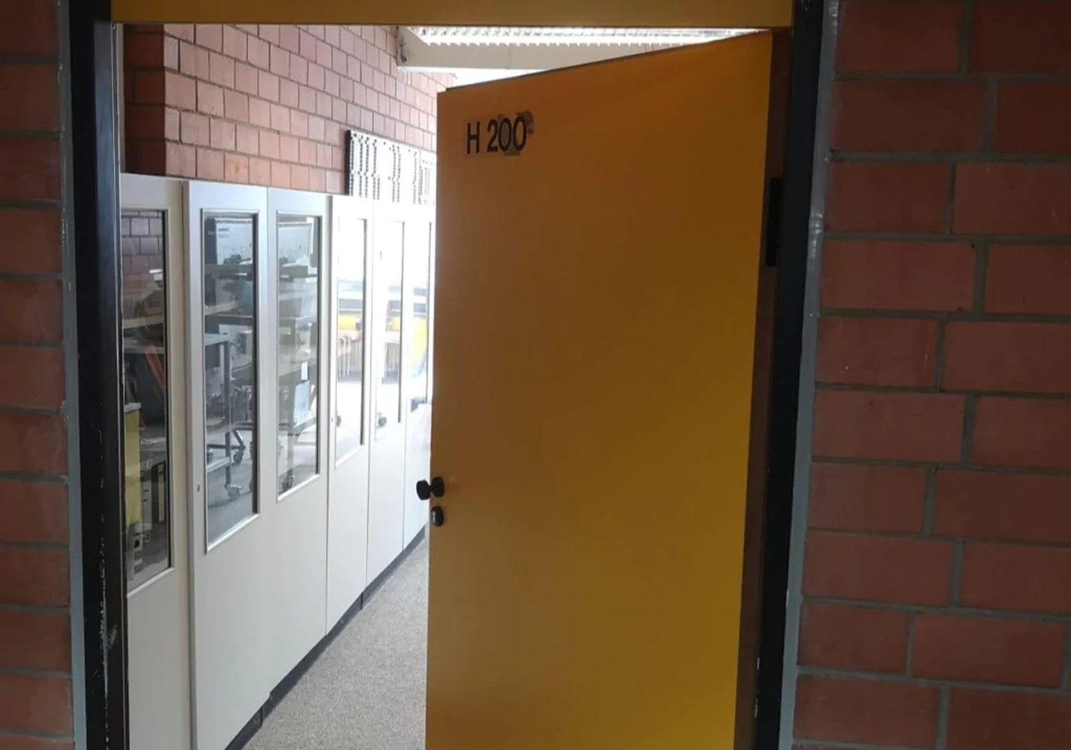 Fugentechnik Buchen - Bau - Teilweise geöffnete gelbe Tür mit der Aufschrift H 200, die zu einem Flur mit weißen Schränken und Backsteinwänden führt.