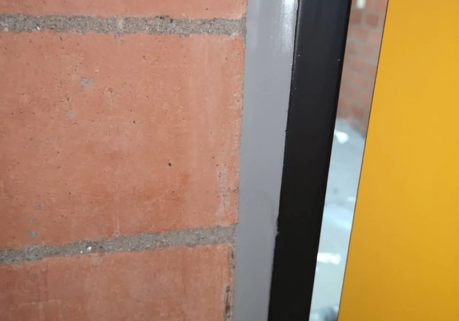 Fugentechnik Buchen - Bau - Nahaufnahme einer roten Backsteinmauer neben einem Türrahmen mit grau-schwarzer Zierleiste und gelbem Türblatt