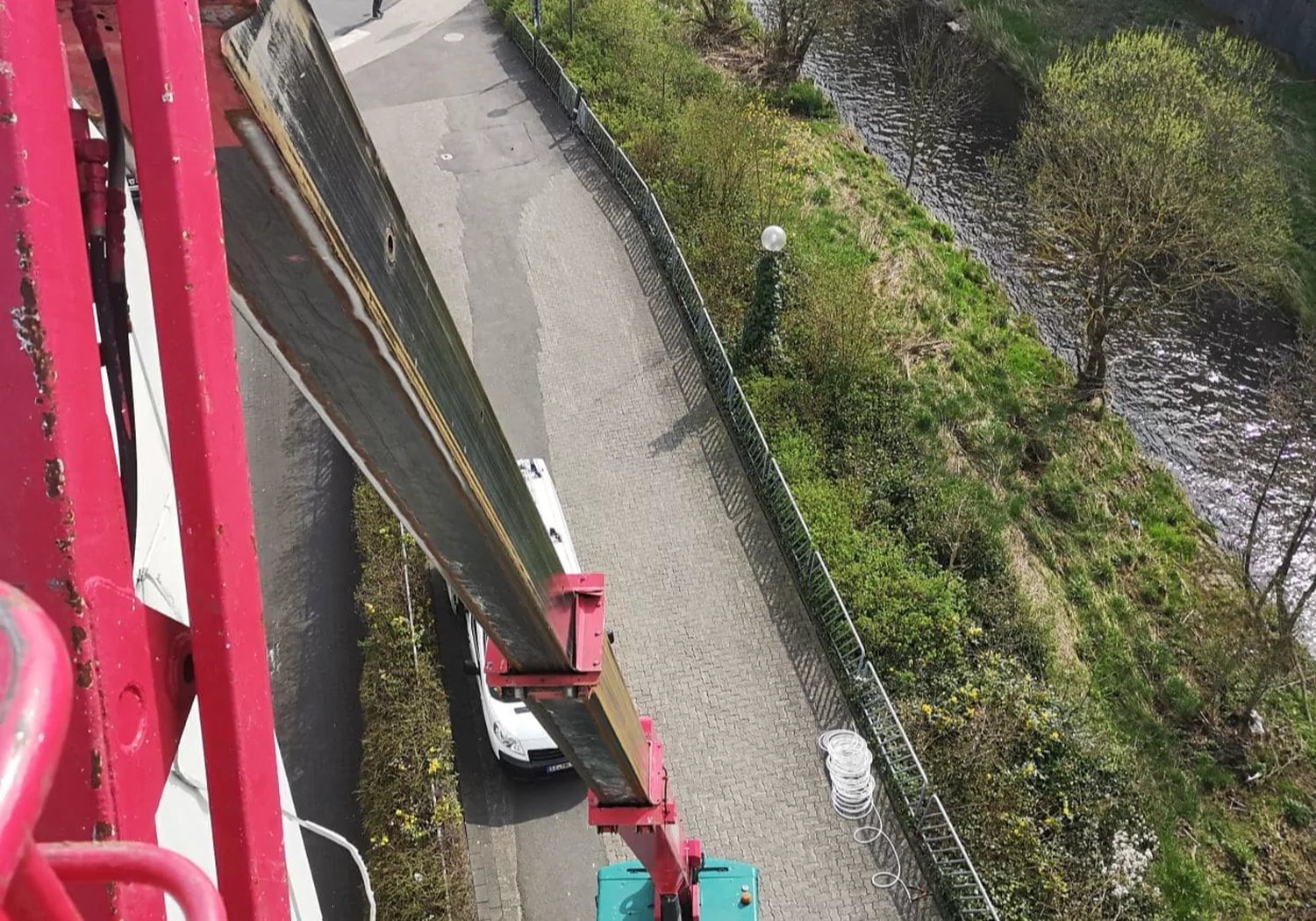 Fugentechnik Buchen - Bau - Blick von oben auf einen roten Kranarm, der sich auf einen weißen Lieferwagen erstreckt, der auf einer asphaltierten Straße neben einer grünen Rasenfläche mit einem Fluss geparkt ist