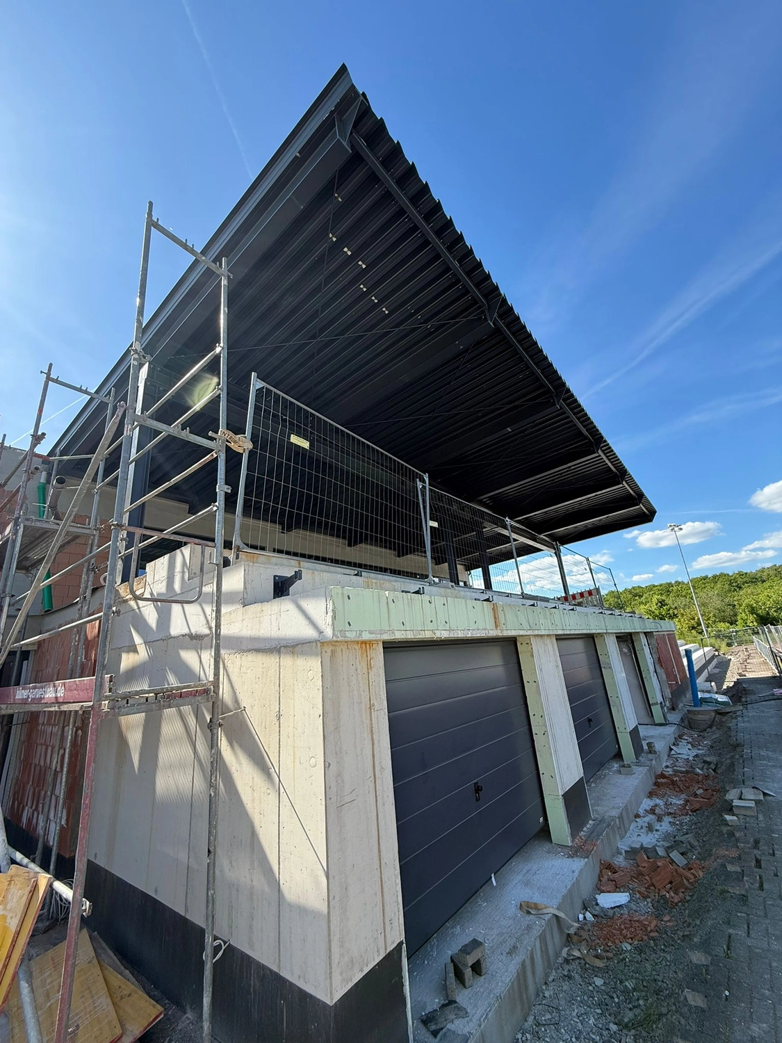 Fugentechnik Buchen – Bau – Baustelle mit einem Gebäude mit drei geschlossenen, dunklen Garagentoren, Gerüst und einem schwarzen Metalldach unter klarem, blauem Himmel