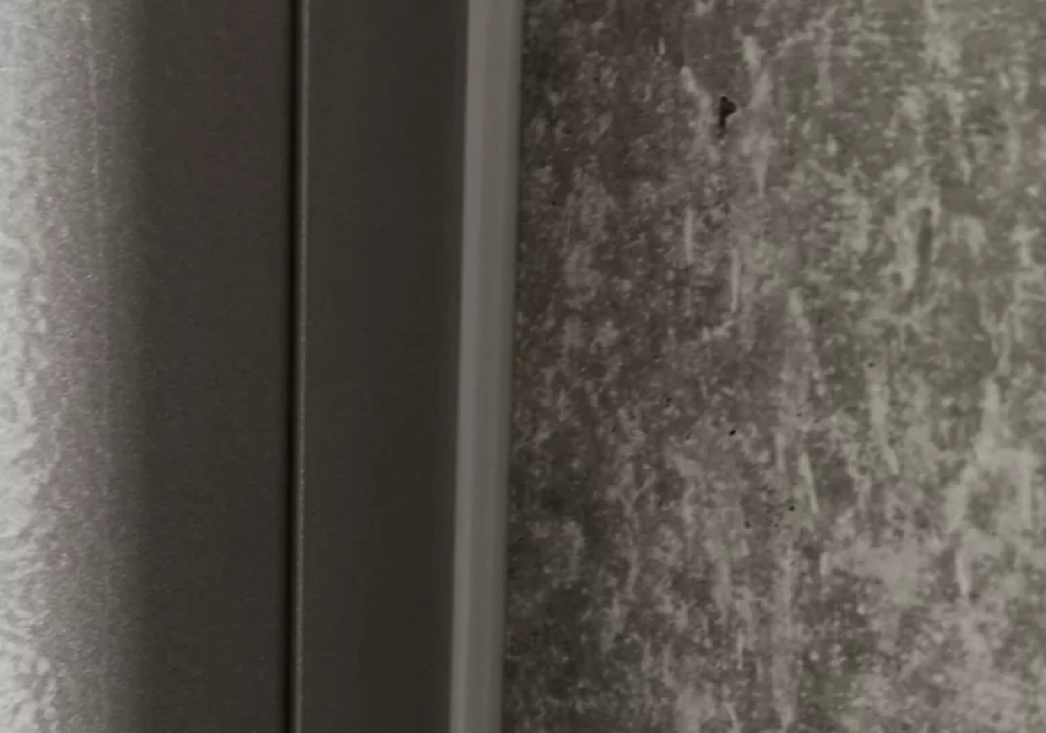 Fugentechnik Buchen - Bau - Nahaufnahme einer Ecke, wo eine glatte, dunkle Metalloberfläche auf eine raue, gesprenkelte, graue Betonwand trifft